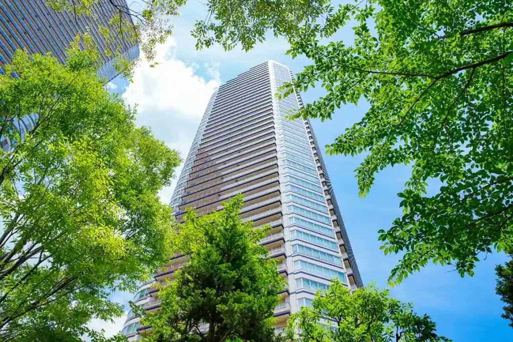 青空を背景に、緑の木々に囲まれた高層マンションを見上げる