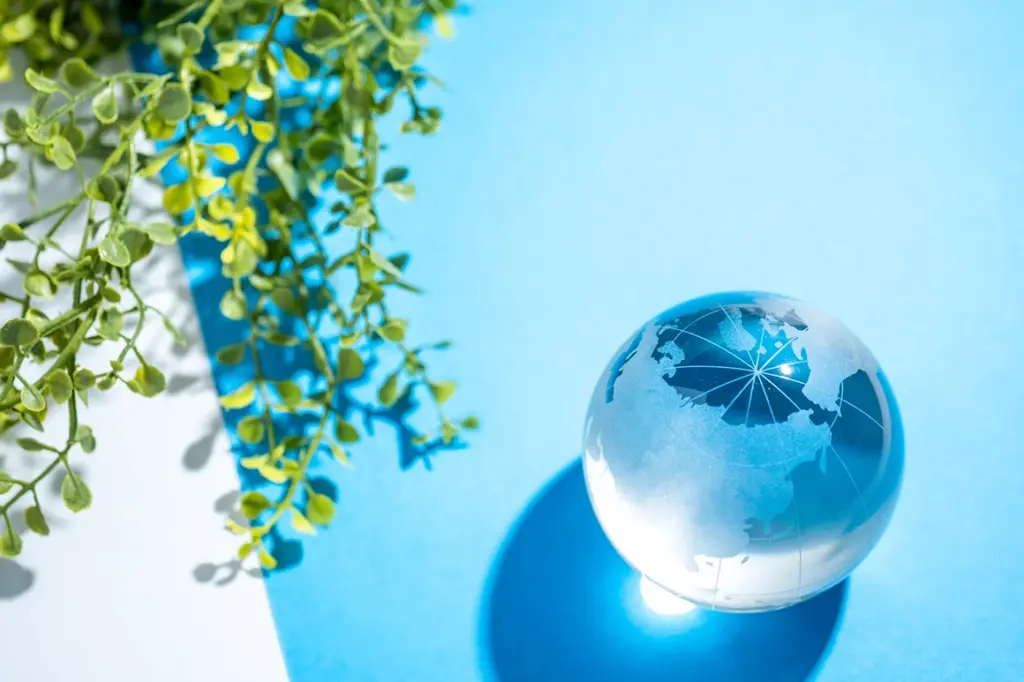 水色の背景に置かれたガラス製の地球儀と、左上から垂れ下がる緑の葉
